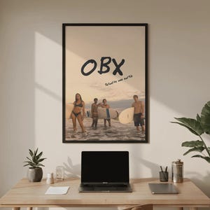May include: A rectangular wall poster in a black frame featuring a photograph of four people standing in the ocean with surfboards at sunset. The text OBX is printed in large black letters above the text Paradise one earth in smaller script.