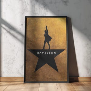 Könnte beinhalten: Gerahmtes Poster mit einer schwarzen Silhouette einer Figur auf einem Stern, darunter das Wort "HAMILTON". Der Hintergrund ist goldfarben strukturiert. Das Poster befindet sich in einem schwarzen Rahmen und lehnt an einer weißen Wand.