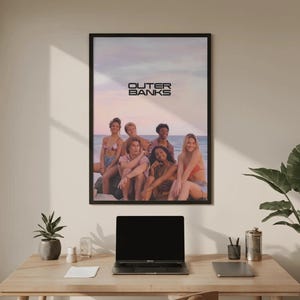 May include: A rectangular wall poster in a black frame featuring the text OUTER BANKS above a group of six people sitting on a beach at sunset. The scene shows a soft sky with pink and blue hues, with the group positioned near the water's edge.