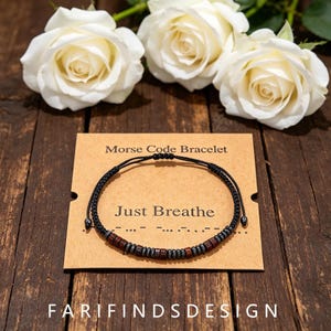Könnte beinhalten: Ein schwarzes Schnurarmband mit kleinen dunkelbraunen Holzperlen und metallisch grauen Perlen in einem Morsecode-Muster. Das Armband liegt auf einer beigen Karte mit dem Aufdruck Morse Code Bracelet und Just Breathe.