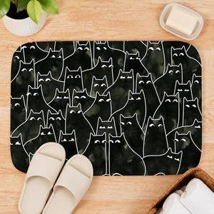 May include: A rectangular bath mat featuring a repeating pattern of black cats with white outlines and simple white eyes. The mat has rounded corners and is positioned on a light wood floor next to a pair of beige slide sandals and a white soap dish.