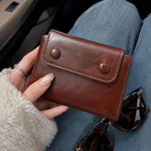 May include: A dark brown leather wallet featuring a rectangular flap secured by two matching brown circular snap buttons. The wallet has a smooth, polished finish and a compact, folded design, showcasing a clean and simple aesthetic for daily use.
