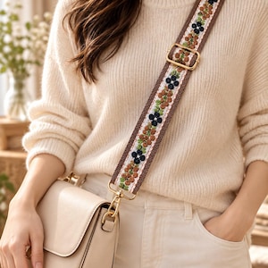 Puede incluir: Una correa para bolso cruzado con base tejida en color marrón y un patrón floral repetitivo en azul marino, tostado y verde. La correa incluye herrajes metálicos en tono dorado, con un deslizador ajustable y un mosquetón unido a un bolso beige.