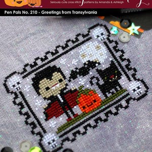 May include: Cross-stitch pattern featuring a vampire, black cat, and jack-o'-lantern. The design is framed with a skull border. The text reads "Pen Pals No. 210 - Greetings from Transylvania" and "The Frosted Pumpkin Stitchery".