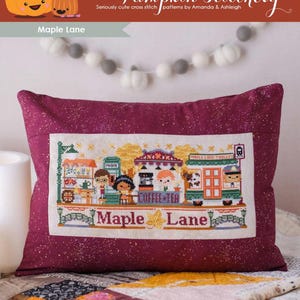 May include: A burgundy pillow with a cross-stitch design featuring a street scene with the text "Maple Lane." The design includes shops, people, and the text "Coffee Tea." The top of the image has the text "The Frosted Pumpkin Stitchery."