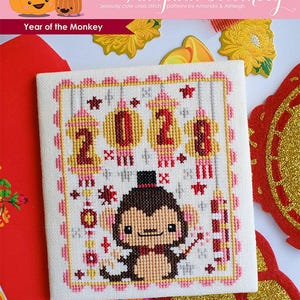 Peut inclure: Un motif de point de croix représentant un singe de dessin animé mignon portant un chapeau haut de forme et un nœud papillon. Le singe est entouré de lanternes rouges et dorées avec l'année 2023 brodée au centre. Le motif est intitulé "Année du singe" et fait partie de la collection "The Frosted Pumpkin Stitchery".