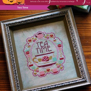 Puede incluir: Un muestrario de punto de cruz enmarcado con las palabras "TEA TIME" en el centro. El diseño presenta una taza de té, teteras y elementos decorativos en rosa, rojo y turquesa. El marco es plateado y el fondo es de tela verde claro.