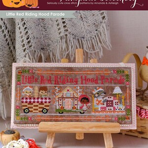 May include: A cross stitch pattern of Little Red Riding Hood Parade, featuring a red hooded girl, a wolf, and a house. The pattern is stitched on a brown fabric with a pink border. The pattern is displayed on a wooden easel.