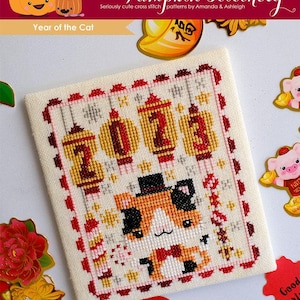 May include: A cross stitch pattern for the Year of the Cat, featuring a cute cartoon cat wearing a top hat and bow tie. The pattern includes the Chinese characters for "2023" and a red and gold color scheme.