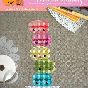 Op de afbeelding: Een kruissteekpatroon voor een scheve toren van macarons. De macarons zijn op elkaar gestapeld en hebben schattige gezichtjes. Het patroon heet "Leaning Tower of Macarons" en is van "The Frosted Pumpkin Stitchery".