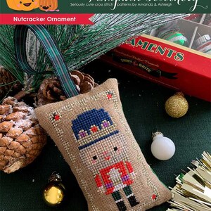 May include: A cross stitch pattern for a nutcracker ornament. The pattern is on a beige fabric with a brown border. The nutcracker is wearing a blue hat, red jacket, and white trousers. The pattern is titled "Nutcracker Ornament".