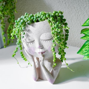May include: A white, face-shaped planter with closed eyes and a finger touching the lips. The planter has pink accents on the cheeks and lips. Green string of pearls plant spills over the top of the planter, creating a unique decorative piece.