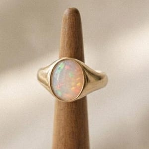 Könnte beinhalten: Ein goldener Siegelring mit einem glatten, ovalen Opal-Edelstein in der Mitte. Der Opal zeigt irisierende grüne, orangefarbene und rosafarbene Lichtreflexe auf einer milchig-weißen Basis. Die Ringschiene besteht aus poliertem Gold mit zulaufender Form.