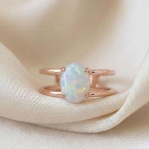 May include: Rose gold ring with an oval opal gemstone. The ring has a split shank design, with two parallel bands that connect to the opal. The opal has a milky white base with flashes of iridescent colour. The ring is set against a soft, cream-coloured fabric.