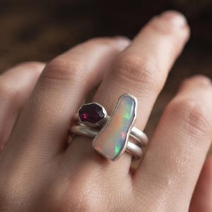 May include: A silver ring set featuring two stacked bands. One band holds a small, faceted deep red garnet stone, while the other band displays a large, elongated, irregular opal with iridescent flashes of green, orange, and yellow light.