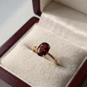 Könnte beinhalten: Ein Goldring mit einem facettierten, oval geschliffenen, tiefroten Granat-Edelstein, der von vier Krappen gehalten wird. Der Ring befindet sich in einer dunkelbraunen Schmuckschatulle mit cremefarbenem Samtfutter, die das polierte Metallband zeigt.