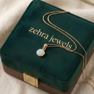 May include: A gold necklace with a round opal pendant rests on a dark green velvet jewelry box. The box has the words "zehra jewels" in gold lettering. The necklace chain is delicate and the pendant is a light, iridescent color.