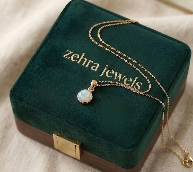 May include: A gold necklace with a round opal pendant rests on a dark green velvet jewelry box. The box has the words "zehra jewels" in gold lettering. The necklace chain is delicate and the pendant is a light, iridescent color.