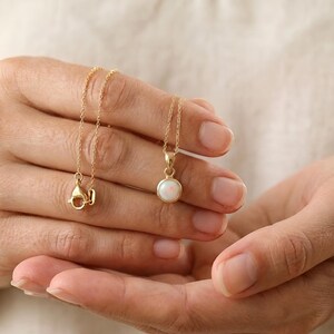 May include: A delicate gold necklace featuring a small, round opal pendant. The opal has a milky white color with subtle iridescence. The necklace is held in a person's hands, showcasing the fine chain and clasp.