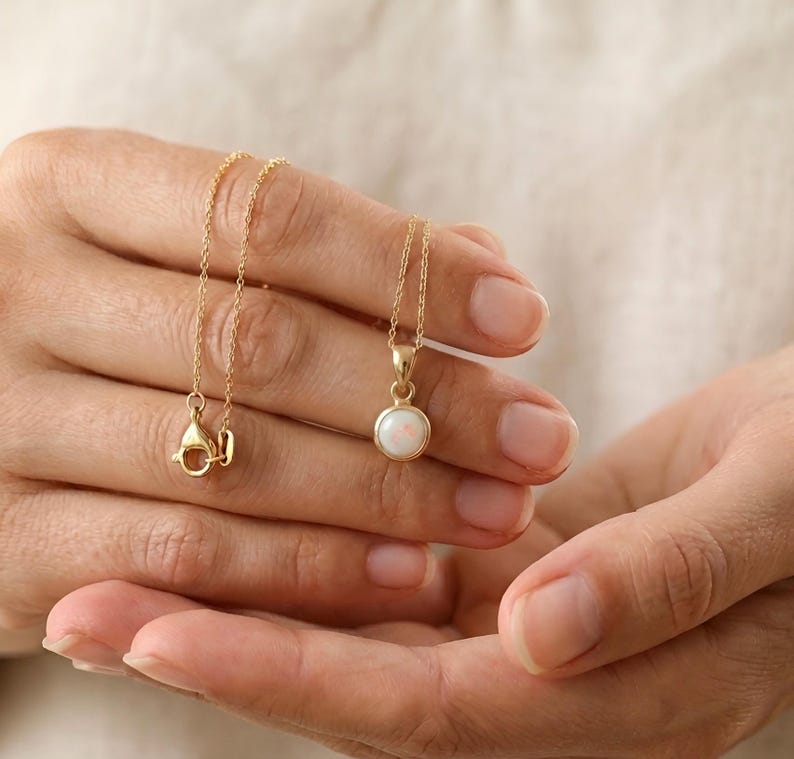 May include: A delicate gold necklace featuring a small, round opal pendant. The opal has a milky white color with subtle iridescence. The necklace is held in a person's hands, showcasing the fine chain and clasp.