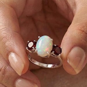 Könnte beinhalten: Ein Silberring mit einem zentralen, ovalen weißen Opal mit irisierenden Farbblitzen, flankiert von zwei runden, tiefroten Granat-Edelsteinen. Die Steine sind in einer Krappenfassung auf einem polierten Silberring mit glatter Oberfläche gefasst.