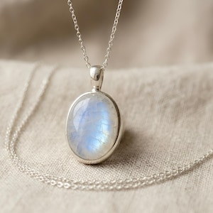 May include: A silver necklace featuring an oval moonstone pendant. The moonstone has a blue and white iridescent glow. The chain is delicate and hangs on a neutral-colored fabric background. The pendant is approximately 1 inch in length.