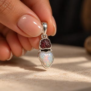May include: A silver pendant featuring a raw, deep red garnet stone positioned above a smooth, heart-shaped white opal with iridescent flashes of color. The pendant includes a silver bail at the top for attachment to a chain or necklace.
