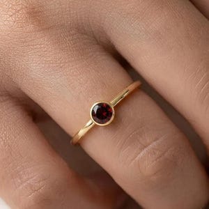 May include: A gold ring featuring a single, round, deep red garnet gemstone set in a smooth bezel setting. The band is thin, polished, and circular, showcasing a minimalist design that highlights the contrast between the gold and the dark red stone.