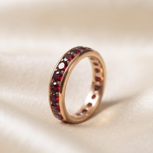 May include: A rose gold eternity band featuring a continuous row of round-cut, deep red garnet gemstones set within the metal channel. The ring has a polished finish and a uniform design that encircles the entire circumference of the band.