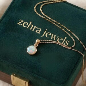 May include: A gold necklace with a round opal pendant rests on a dark green velvet jewelry box. The box is embossed with the words "zehra jewels" in gold lettering. The opal pendant is white with iridescent colors.