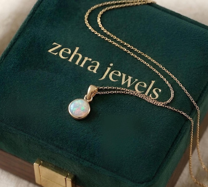 May include: A gold necklace with a round opal pendant rests on a dark green velvet jewelry box. The box is embossed with the words "zehra jewels" in gold lettering. The opal pendant is white with iridescent colors.