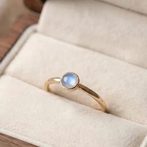 Può includere: Un anello in oro con una singola pietra di luna rotonda a cabochon incastonata a lunetta. La gemma mostra un morbido riflesso blu iridescente sulla fascia in oro lucido, che presenta un profilo liscio e arrotondato dal design semplice.