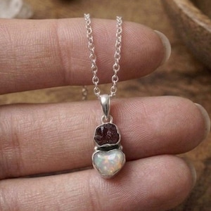 Può includere: Una collana con pendente in argento caratterizzata da una piccola pietra granato grezza rosso scuro posizionata sopra una pietra opale bianca iridescente e liscia. Le due pietre sono incastonate in una montatura d'argento e una catenina.