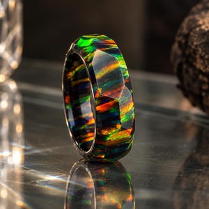Op de afbeelding: Een gefacetteerde ring met een donkere basis en levendige, iriserende strepen van groen, oranje en rood over het hele oppervlak. De band heeft een geometrische, veelzijdige buitenafwerking die het licht reflecteert en kleurspelingen vertoont.