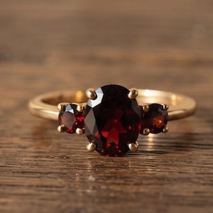 Op de afbeelding: Een gouden ring met een grote, ovaal geslepen dieprode granaatsteen in het midden, geflankeerd door twee kleinere, rond geslepen rode granaatstenen. De stenen worden op hun plaats gehouden door gouden pootjes op een gladde, gepolijste gouden ring.