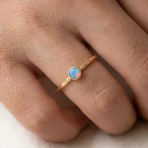 May include: A delicate gold ring featuring a small, round opal stone. The opal displays a mix of blue, white, and peach hues. The ring is shown on a finger, highlighting the minimalist design and the gemstone's subtle colors.