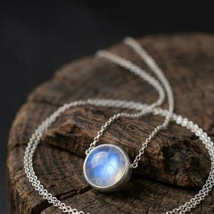 May include: A silver necklace featuring a large, round, iridescent blue gemstone pendant. The pendant is set in a silver bezel and hangs from a delicate silver chain. The necklace rests on a textured, dark brown wooden surface.