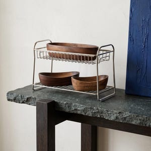 Peut inclure: Un présentoir métallique à deux niveaux contenant trois bols en bois. Les bols ovales sont de couleur marron foncé. Le présentoir argenté est posé sur une surface en pierre grise foncée. Une peinture abstraite bleu foncé est en arrière-plan.