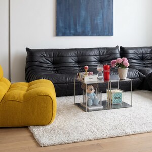 May include: A modern living room with a black leather sofa, a yellow armchair, and a small, square, metal coffee table. A large blue abstract painting hangs on the white wall. The room has a white rug and a floor lamp.