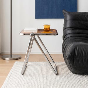 May include: A modern side table with a transparent amber-colored top and a polished chrome frame. A stack of books and a small amber glass sit on the table. The table is on a white rug.