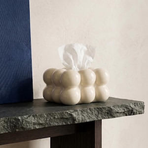 May include: A cream-colored tissue box with a unique, rounded design. The box is filled with white tissues, with some tissues pulled out. The tissue box sits on a dark gray stone surface, with a dark brown wooden table leg visible.