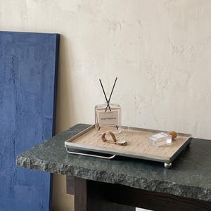 May include: A square glass bottle with black reeds and the text "Mose Fragrance" sits on a decorative tray with a watch and perfume bottle. The tray has a silver frame and is on a dark green stone table.