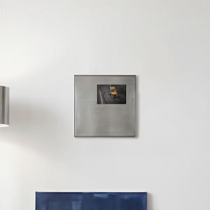 Op de afbeelding: Een vierkant wandkunstobject van geborsteld metaal met een gecentreerde, rechthoekige fotoprint. De foto toont een enkele, felgele stoel op een grijs, geplaveid oppervlak, wat een scherp contrast vormt met het metalen frame van het kunstwerk.