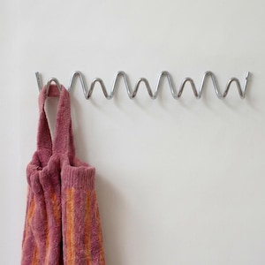 May include: A silver-colored metal wall-mounted coat rack featuring a continuous, repeating zigzag or wave-like design. A textured, striped fabric bag in shades of dusty rose and orange hangs from one of the loops of the metal rack.