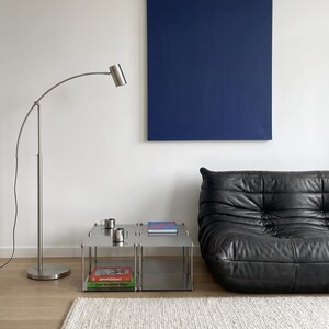 May include: A modern living room scene featuring a black leather sofa, a stainless steel side table with books and mugs, and a blue canvas art piece. A silver arc floor lamp and a white rug complete the minimalist decor.