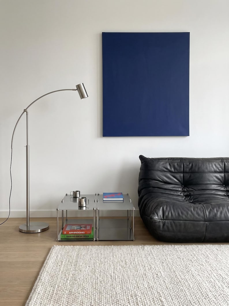 May include: A modern living room scene featuring a black leather sofa, a stainless steel side table with books and mugs, and a blue canvas art piece. A silver arc floor lamp and a white rug complete the minimalist decor.