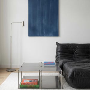 May include: A modern living room featuring a stainless steel coffee table with books, a black leather sofa, a blue abstract painting, and a silver floor lamp. The room has a minimalist design with a white rug.