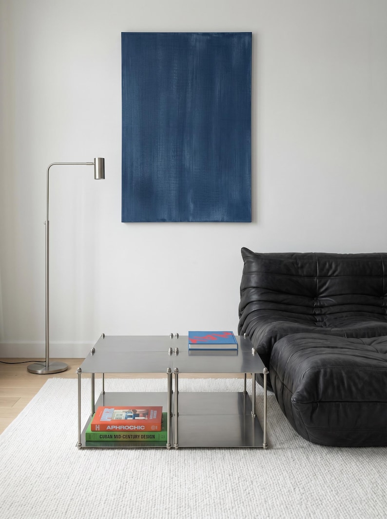 May include: A modern living room featuring a stainless steel coffee table with books, a black leather sofa, a blue abstract painting, and a silver floor lamp. The room has a minimalist design with a white rug.