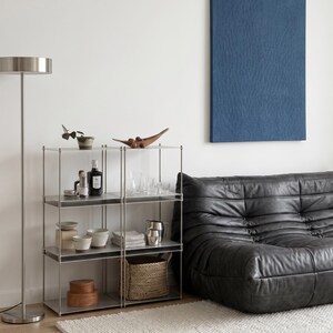 May include: A modern living room scene featuring a metal shelving unit with various decorative items, including bowls, a wooden bowl, and bottles. A black leather sofa and a blue wall art piece are also present, along with a floor lamp and a white rug.