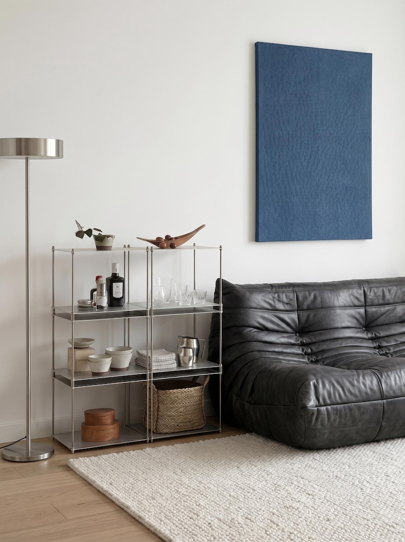 May include: A modern living room scene featuring a metal shelving unit with various decorative items, including bowls, a wooden bowl, and bottles. A black leather sofa and a blue wall art piece are also present, along with a floor lamp and a white rug.